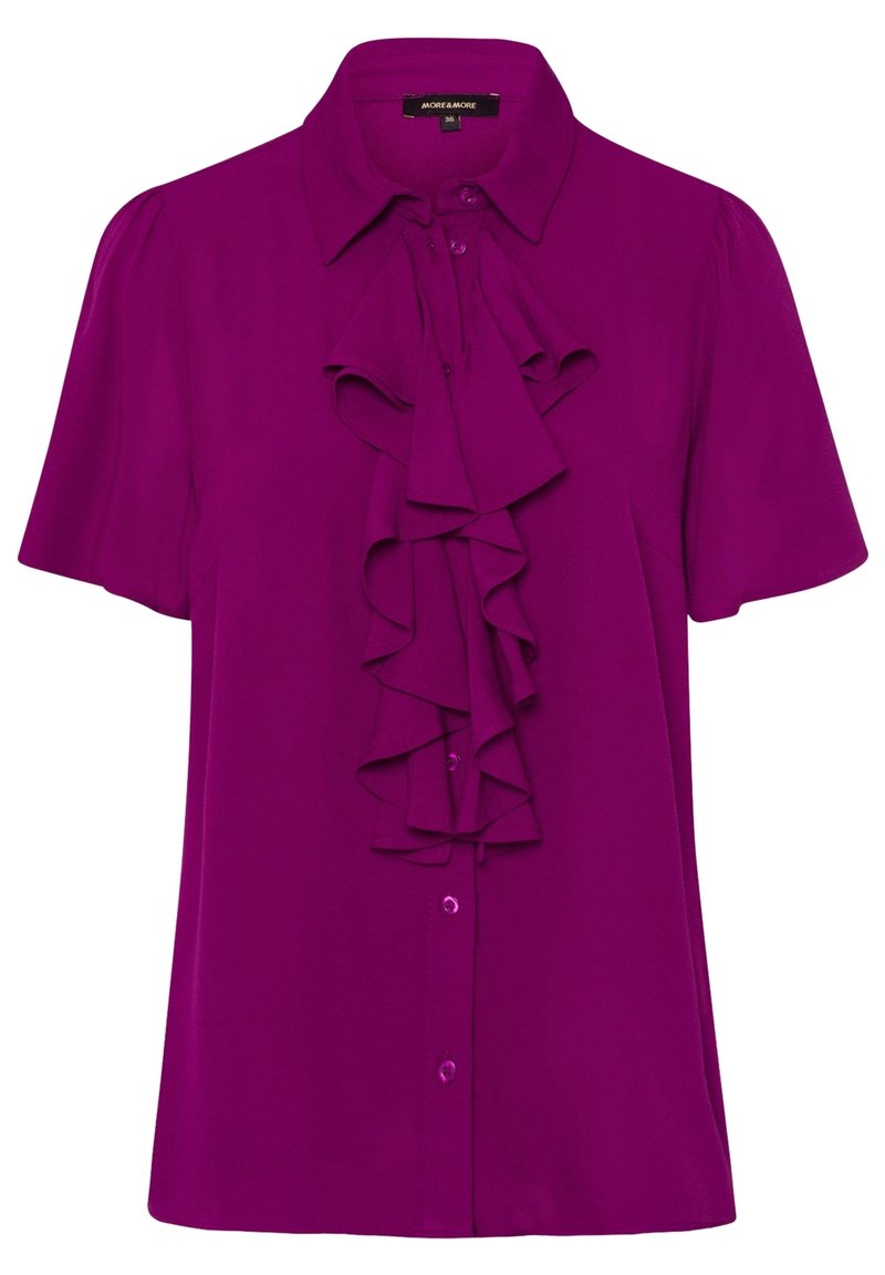 Bluzka z krótkim rękawem w jasnym fioletowym kolorze, z kołnierzykiem i falbanami z przodu. Posiada zapięcie na guziki oraz gładki, lekki materiał.