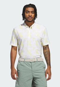 Short-sleeve polo in white with yellow abstract pattern; features a collar, three-button placket, and logo on the chest; paired with mint green shorts.