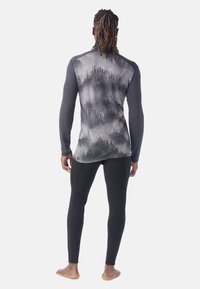 Persoon met dreadlocks die een nauwsluitende lange mouw top draagt met een zwart-grijs bospatroon en een paarse  zwarte leggings, staande op blote voeten.