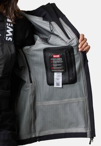 Musta takki, jossa harmaa vuori, vetoketjullinen verkkotasku RECCO-teknologialla ja erilaisia sisätaskuja.