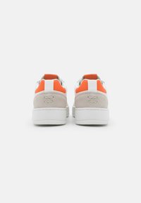 Białe sneakersy o solidnej konstrukcji, z pomarańczowymi i białymi akcentami z tyłu oraz teksturowaną panelą z beżowego zamszu.