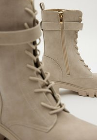 Botas de tobillo beige con cordones, cremallera lateral, suela gruesa y detalle de correa, fotografiadas sobre un fondo neutro.