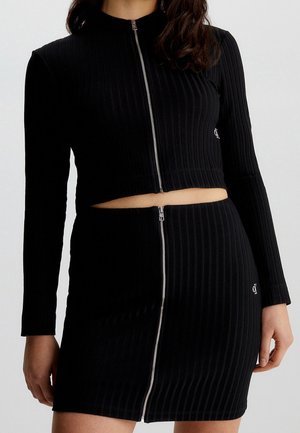 Kobieta w czarnym prążkowanym dwuczęściowym stroju z długimi rękawami i srebrnymi suwakami z przodu, odsłaniającymi brzuch.