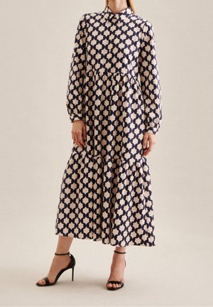 Femme portant une robe midi à manches longues et col avec un motif géométrique bleu marine et beige, associée à des talons hauts noirs à bride à la cheville.