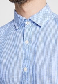 Chemise à rayures bleu clair avec col, boutons blancs et tissu texturé, vue en gros plan de la partie supérieure avant.