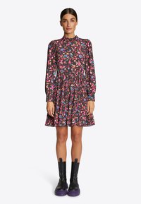 Květinové šaty s dlouhým rukávem na černém pozadí, se spoustou barevných vzorů, vysokým výstřihem a skládací sukní; nošeny s černými a fialovými botami.