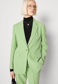 Hellgrüner taillierter Blazer mit einem einzelnen Knopf, Reverskragen und Vorderstaschen. Über einem schwarzen Rollkragenpullover getragen, kombiniert mit schwarzen Accessoires.