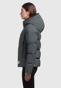 Chaqueta acolchada gris con capucha, que cuenta con un bolsillo con cremallera, textura esponjosa y una etiqueta de logo en el dobladillo. El diseño es ceñido y moderno.