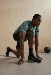 Black athletic shoes with green accents, lightweight fabric, and textured upper; model in a crouched position wearing a green shirt and black shorts.