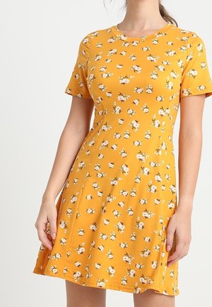 Żółta musztardowa sukienka z krótkim rękawem, z drobnym białym kwiatowym wzorem, dopasowaną talią i rozkloszowanym dołem, noszona przez osobę z rękami spoczywającymi po bokach.