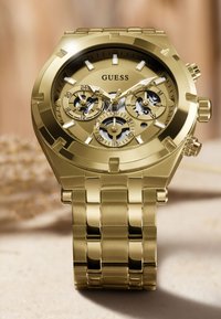 Złoto-tonowany zegarek ze stali nierdzewnej z trzema subtarczami, dużymi cyfrowymi znacznikami i teksturowaną tarczą. Posiada masywny bransoletę z ogniwami.