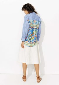 Chemise à manches longues rayée bleu et blanc avec un imprimé floral coloré dans le dos, associée à une jupe blanche et des sandales marron.
