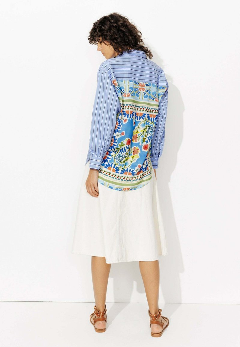 Chemise à manches longues rayée bleu et blanc avec un imprimé floral coloré dans le dos, associée à une jupe blanche et des sandales marron.