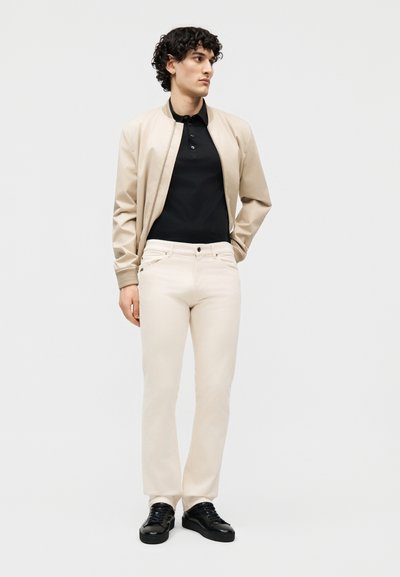 Άνδρας που φοράει μπεζ μπουφάν τύπου bomber, μαύρο πόλο πουκάμισο, ανοιχτόχρωμο παντελόνι και μαύρα αθλητικά παπούτσια, στέκεται μπροστά σε μονόχρωμο γκρι φόντο.