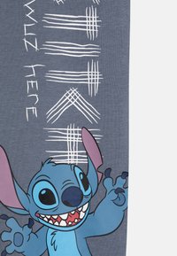 Grauer Stoff mit einem blauen Cartoon-Charakter mit großen Ohren und einem breiten Grinsen, umgeben von weißen geometrischen Linien und dem Text "Wuz Here."