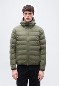 Νεαρός με σγουρά μαλλιά που φοράει καφέ-ελιά μπουφάν με ψηλό γιακά και μαύρο παντελόνι, στέκεται μπροστά σε απλό φόντο.