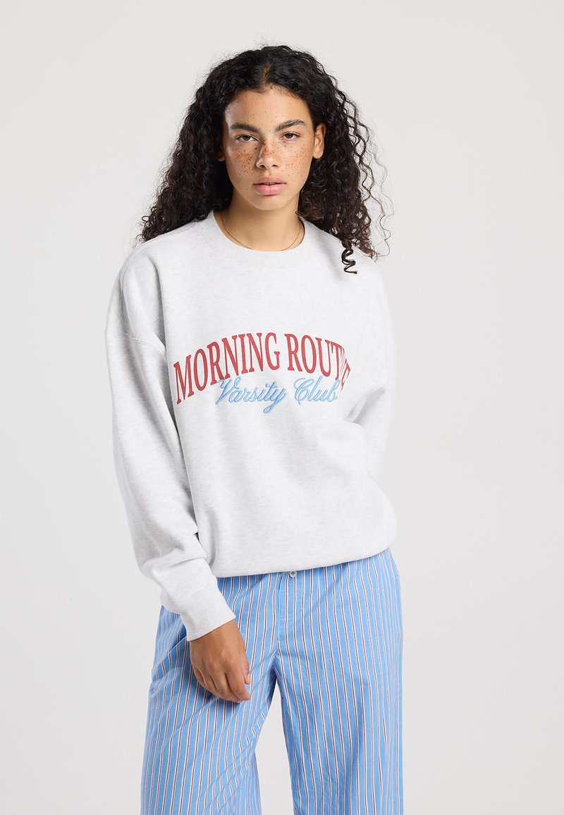 Grijze sweatshirt met rode en blauwe tekst "MORNING ROUTE Varsity Club." Gepaarde met blauw gestreepte broek. Zacht, casual ontwerp.