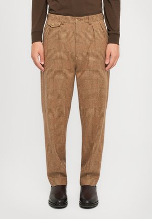 Man wearing brown plaid tailored trousers with side flap pocket and dark brown leather boots, standing on white background.