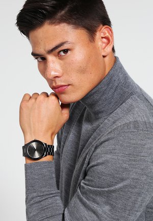 Joven con jersey de cuello alto gris apoyando la barbilla en el puño, lleva un reloj de pulsera metálico negro con esfera degradada y correa de eslabones metálicos.