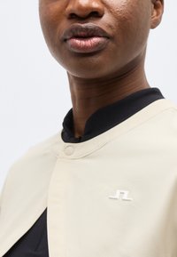 Közeli kép egy személyről, aki világos bézs színű dzsekit visel kerek nyakkal és egy kis fehér, geometriai kitűzővel a mellkasán.