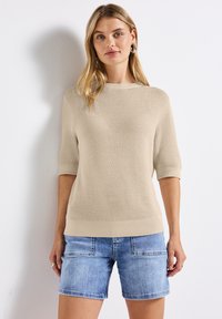 Beige neulepusero, jossa on lyhyet hihat ja ribbireunat, yhdistettynä vaaleansinihin denimitseihin, joissa on etutaskut ja rento istuvuus.