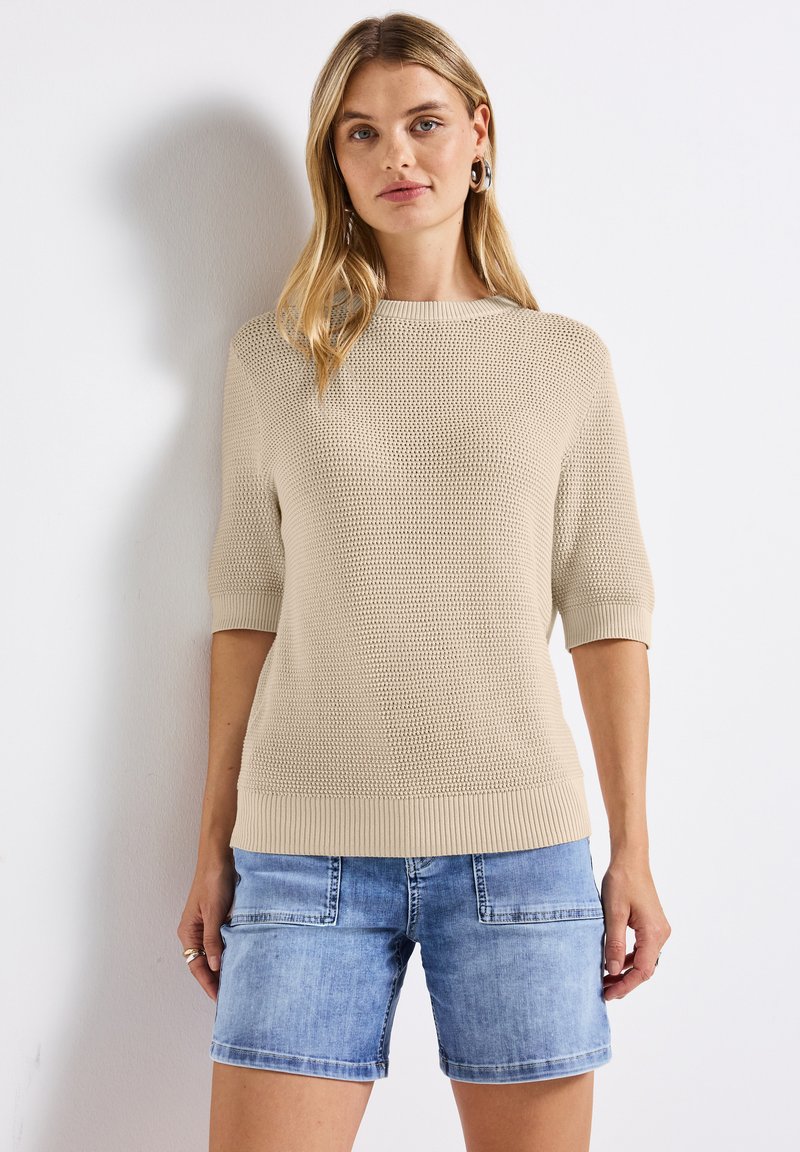 Beige neulepusero, jossa on lyhyet hihat ja ribbireunat, yhdistettynä vaaleansinihin denimitseihin, joissa on etutaskut ja rento istuvuus.