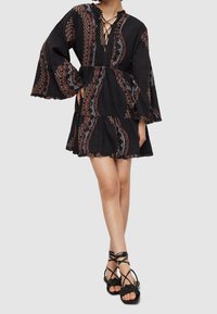 Femme portant une mini-robe noire à motifs avec de longues manches évasées et un col lacé, associée à des sandales plates noires à brides.