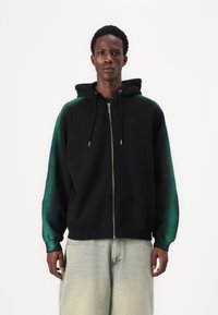 Svart zip-up hettegenser med grønne gradientermidler, mykt stoff, justerbar snor i hetta og ribbestrikkede mansjetter. Sammen med lyseblå jeans.