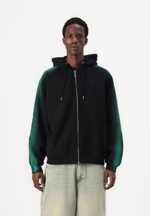 Schwarze Zip-Hoodie mit grün verlaufenden Ärmeln, aus weichem Stoff, mit verstellbarem Kordelzugkapuze und gerippten Bündchen. Kombiniert mit hellblauen Jeans.