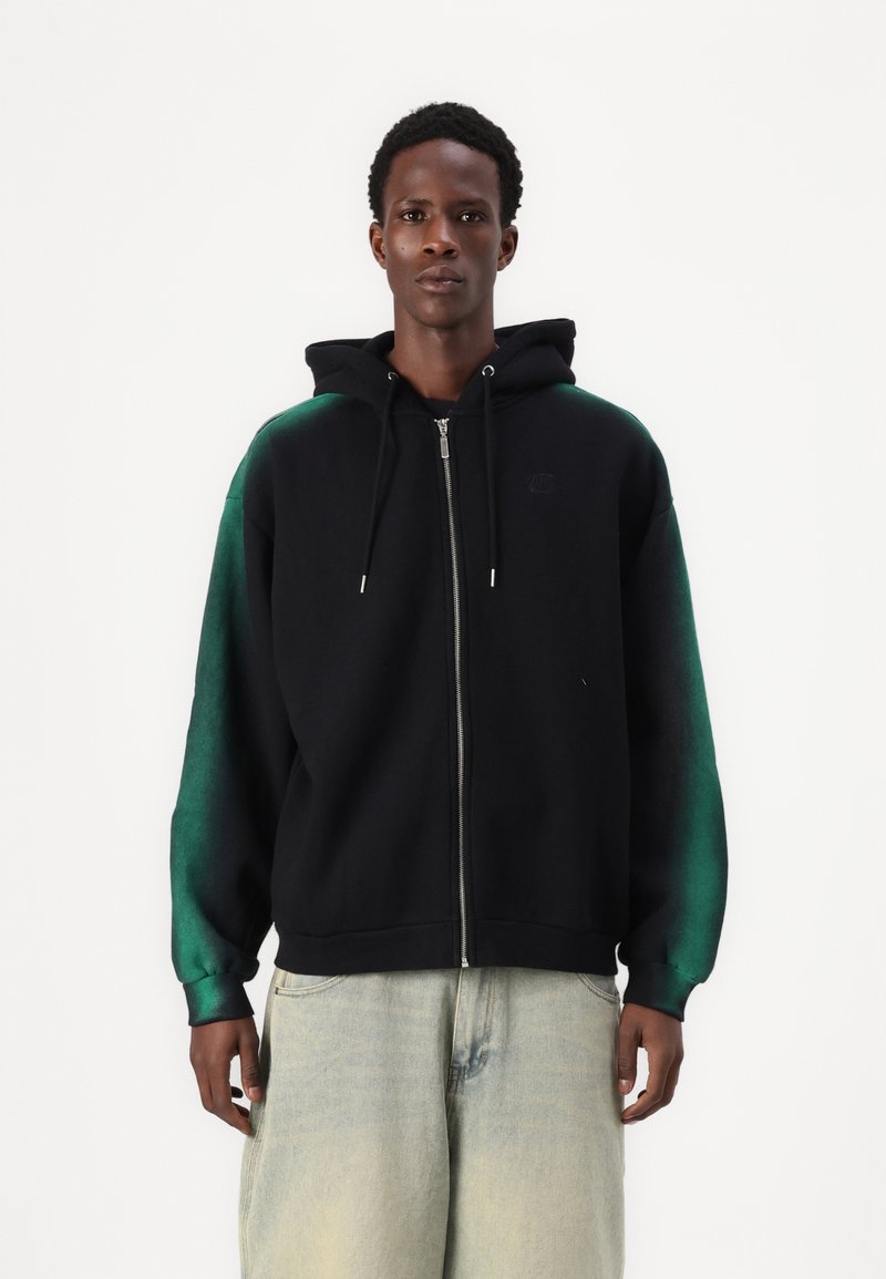 Svart zip-up hettegenser med grønne gradientermidler, mykt stoff, justerbar snor i hetta og ribbestrikkede mansjetter. Sammen med lyseblå jeans.