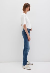 Weißes kurzärmeliges Oberteil, kombiniert mit mitteldunklen Jeans und glänzenden silbernen Schuhen. Das Modell steht seitlich vor einem hellen Hintergrund.