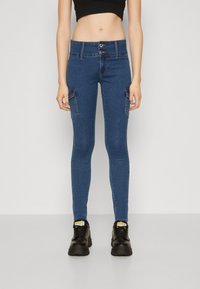 Jeans ajustados de mezclilla azul con bolsillos estilo cargo, cintura alta y cierre de dos botones. Combinados con zapatillas deportivas negras de suela gruesa.