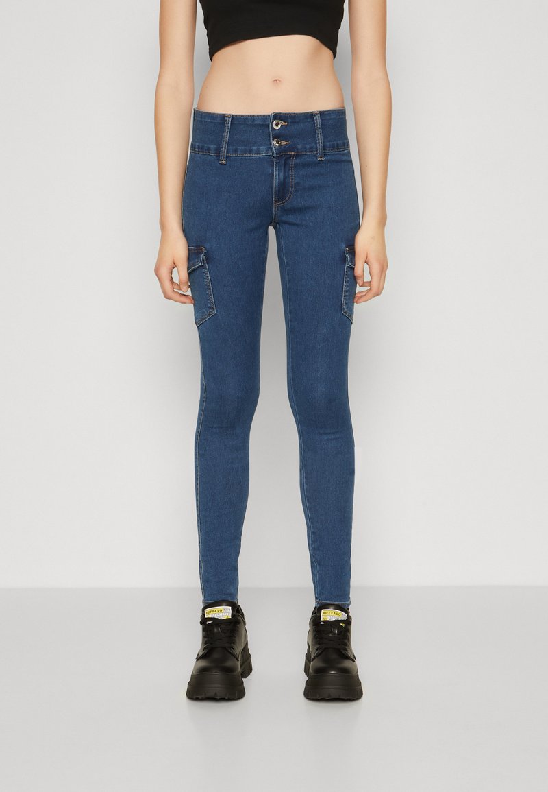 Jeans ajustados de mezclilla azul con bolsillos estilo cargo, cintura alta y cierre de dos botones. Combinados con zapatillas deportivas negras de suela gruesa.