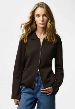 Brązowa kurtka na zamek z kołnierzem, z długimi rękawami i gładką dzianą fakturą. W zestawie z niebieskimi dżinsami.