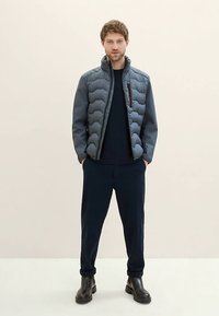 Grå dunjakke med quiltet design, navyblå skjorte under, navyblå bukser og sorte ankelstøvler, sat mod en ensfarvet baggrund.