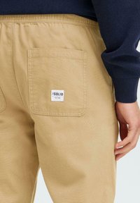 Beige pamut nadrág látható hátsó zsebbel és márkás címkével. Simább felület és rugalmas derékrész.