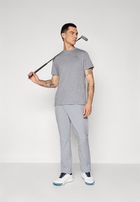 Šedé tričko s krátkým rukávem a kulatým výstřihem, spárované se světle šedými kalhotami. Model drží golfovou hůl a má na sobě bílé golfové boty s modrými akcenty.