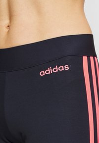 Marina leggings med en tjock midjeband, rosa broderad "adidas"-logotyp och rosa vertikala ränder längs sidorna. Slät, elastisk tyg.