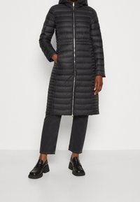 Sort puffet jakke med hætte, vertikalt quiltet design, frontlynlås og to sidelommer, parret med mørke bukser og chunky sko.