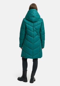 Teal puffer coat with a chevron pattern, angled hem, and a large hood, paired with black jeans and chunky black boots.