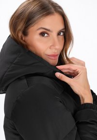 Giacca a vento nera con cappuccio, dotata di texture trapuntata. Il modello tiene delicatamente il colletto. Il tessuto liscio e i polsini elastici creano una vestibilità strutturata.