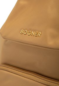 Zaino in nylon beige con tasca frontale con zip, logo "BOGNER" in oro embossato e superficie texturizzata. Linee pulite e design funzionale.