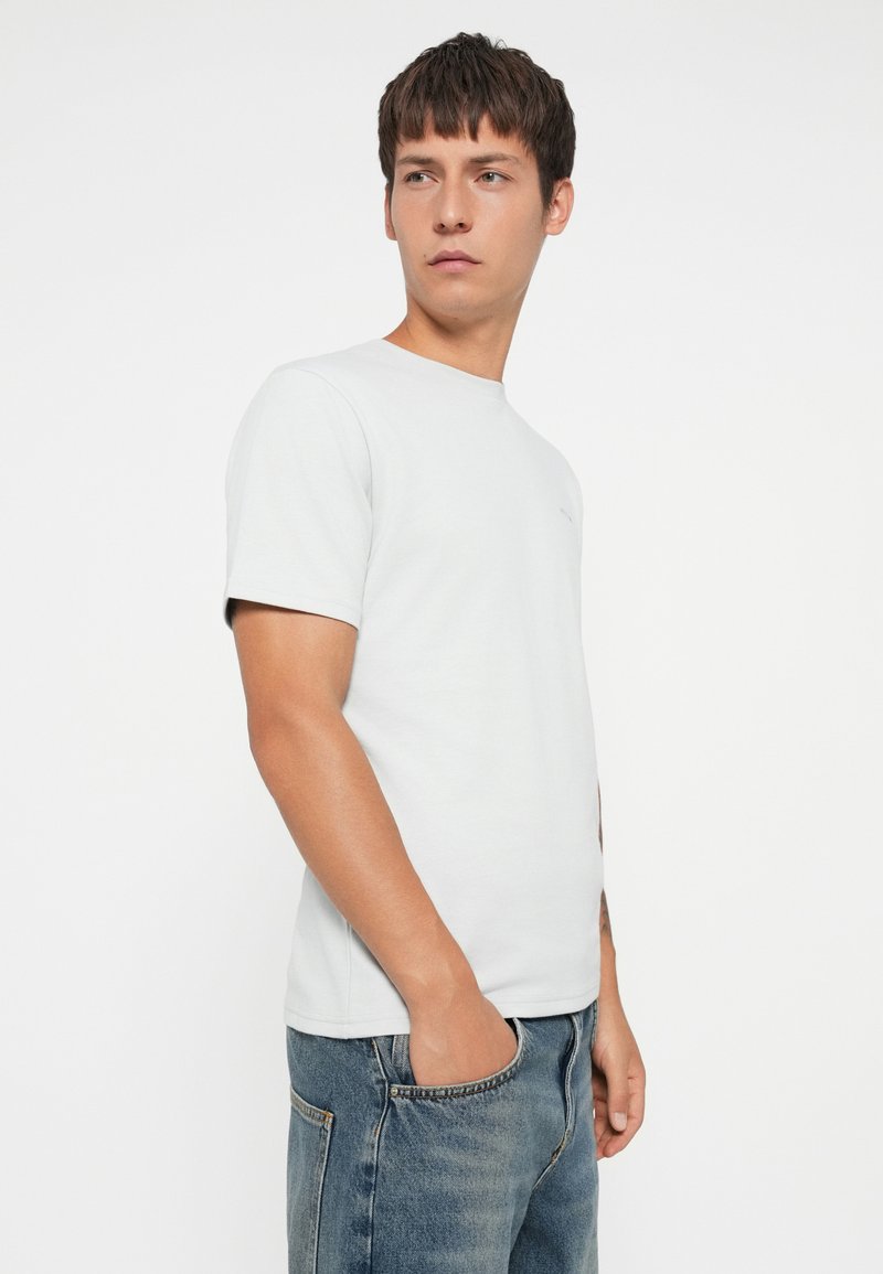 Camiseta de manga corta gris claro, diseño de cuello redondo, fabricada en algodón suave. Llevada con jeans de mezclilla azules, que cuentan con un bolsillo lateral.