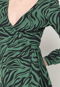 Robe portefeuille verte avec un motif à rayures zèbre noires, encolure en V, manches longues et un détail de ceinture latérale. Confectionnée dans un tissu doux et extensible.