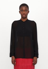 Camicia nera trasparente a maniche lunghe con un frontale bottonato e un pannello plissettato sul petto. Presenta un colletto rotondo e maniche con polsini.