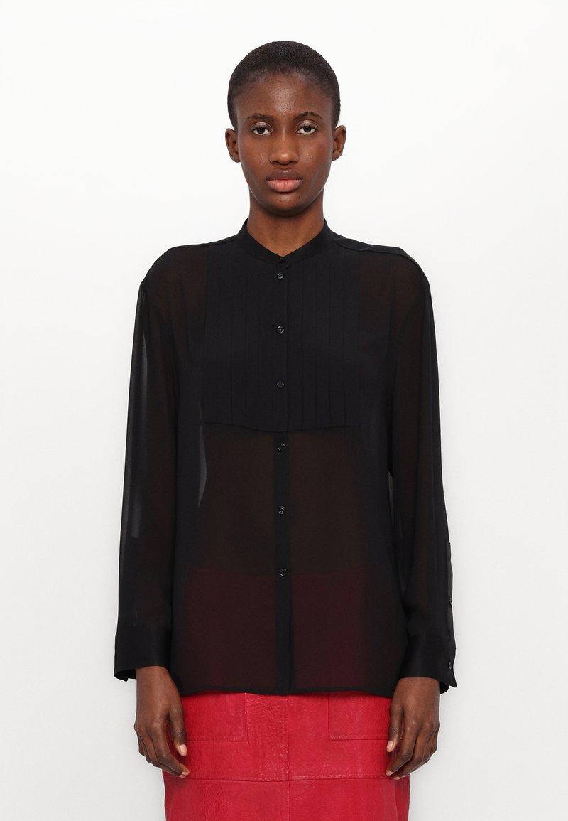 Camicia nera trasparente a maniche lunghe con un frontale bottonato e un pannello plissettato sul petto. Presenta un colletto rotondo e maniche con polsini.