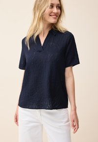 Marineblauwe blouse met korte mouwen, een V-hals en een gestructureerd bladpatroon. De stof is lichtgewicht en heeft een losse pasvorm. Gekleed met witte broek.