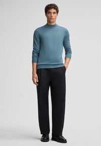 Blauer gestrickter Rollkragenpullover mit gerippten Bündchen, kombiniert mit schwarzen Hosen. Das Model steht mit den Händen in den Taschen und trägt schwarze Schuhe.