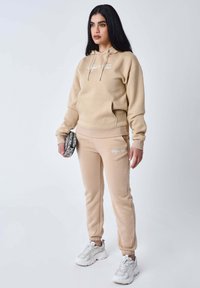Sudadera beige con bolsillo frontal y cordones; joggers a juego con tobillos acampanados. Detalle de logo blanco en ambas piezas. Zapatillas plateadas y bolsa con estampado.