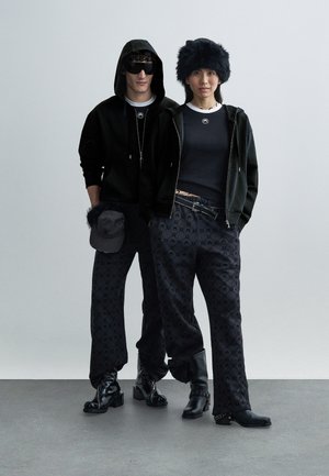 Čierna mikina s kapucňou na zips, skombinovaná s priliehavým čiernym topom a tmavými vzorovanými nohavicami. Doplnky zahŕňajú čiernu čiapku a kožušinovú čiapku. Čierne topánky.